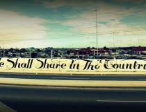 Banner saying "We shall share in the country's wealth" outside shanty town on N2. Photo by Mollie Liesner.