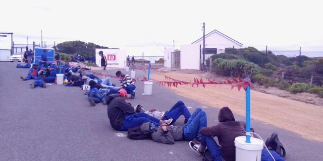 Photo of workers in uniform lying on the pavement outside gates of I&J
